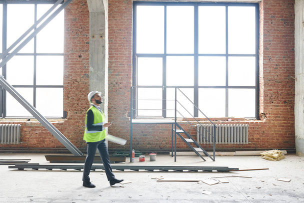 Jonge voorman in uniform lopen in onvoltooide bouw tijdens het controleren van het werk van bouwers - Foto, afbeelding