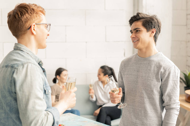 Two happy guys in casualwear discussing curious ideas for toasts while having champagne on background of girls - Foto, imagen