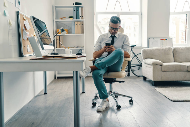 Jeune homme réfléchi en chemise et cravate en utilisant un téléphone intelligent tout en étant assis au bureau
 - Photo, image