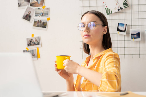 Jovem designer sério do sexo feminino com caneca amarela olhando para você através de óculos enquanto sentado no local de trabalho no escritório
 - Foto, Imagem