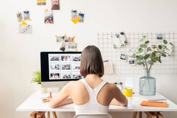 Rückansicht einer jungen Designerin oder Redakteurin in Freizeitkleidung, die vor einem Monitor sitzt, während sie an einer neuen Kollektion von Fotos arbeitet - Foto, Bild