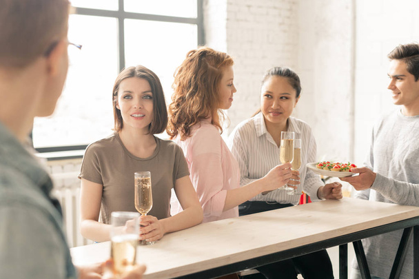 Şampanya flütlü genç kadın masada otururken ve arkadaşları atıştırmalık ve içecekler içerken erkek arkadaşıyla konuşurken - Fotoğraf, Görsel