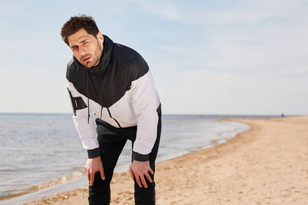 Young Caucasian athlete standing on sandy beach by waterside after tiresome training in the morning - Fotografie, Obrázek