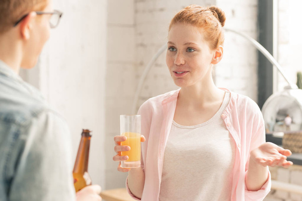 hübsche Ingwerhaarige mit einem Glas Orangensaft, die vor einem Kerl mit einer Flasche Bier steht, während sie auf einer Party plaudert - Foto, Bild