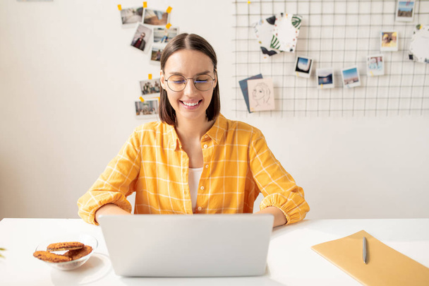 junge fröhliche brünette Frau in Freizeitkleidung sitzt vor dem Laptop, schaut Videos und isst Snacks am Schreibtisch - Foto, Bild
