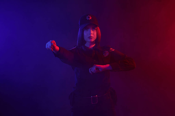 Vörös hajú női rendőr pózol a kamera ellen, fekete háttér, piros és kék háttérvilágítással. - Fotó, kép
