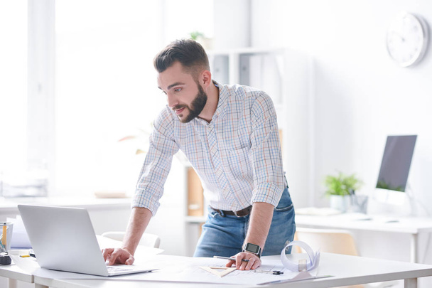 junger selbstbewusster Geschäftsmann oder Architekt in Hemd und Jeans blickt auf Laptop-Display, während er sich am Arbeitstag über Schreibtisch beugt - Foto, Bild