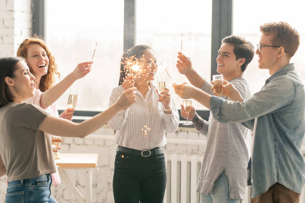 ホームパーティーでライフイベントを祝うシャンパンとベンガルライトのフルートを持つ若い興奮した女の子と男 - 写真・画像