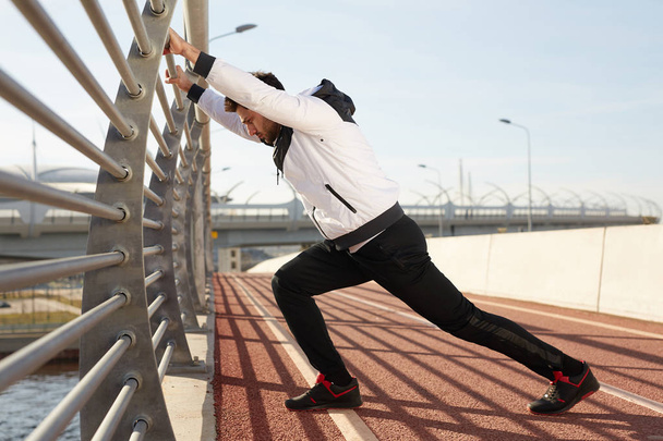 Mladý aktivní sportovec opírající se o zábradlí mostu při cvičení pro nohy na závodní dráze venku - Fotografie, Obrázek