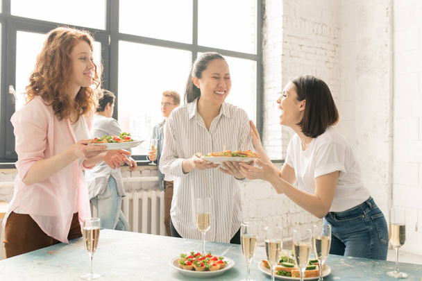 Дві дівчини сміються в повсякденному одязі і їх друг стоїть за столом, подаючи його з закусками для вечірки
 - Фото, зображення