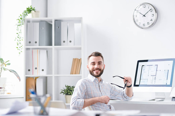 Mladý úspěšný architekt v neformálním oblečení při pohledu na vás, zatímco sedí na pracovišti s počítačovým monitorem na stole - Fotografie, Obrázek