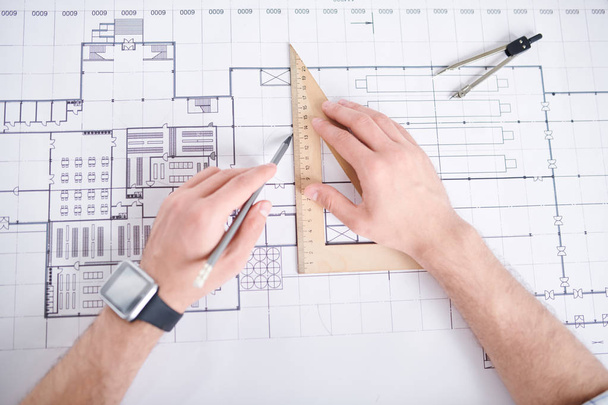 Engineer with pencil drawing straight line by edge of wooden triangle measuring tool while working over sketch of construction - Photo, Image