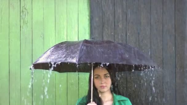 Heavy rain pours on girl sheltered under umbrella. Girl with her hair laid is under the umbrella, heavy rain intensifies and pours heavily. Girl on colored background looks into camera with a light smile. - Footage, Video