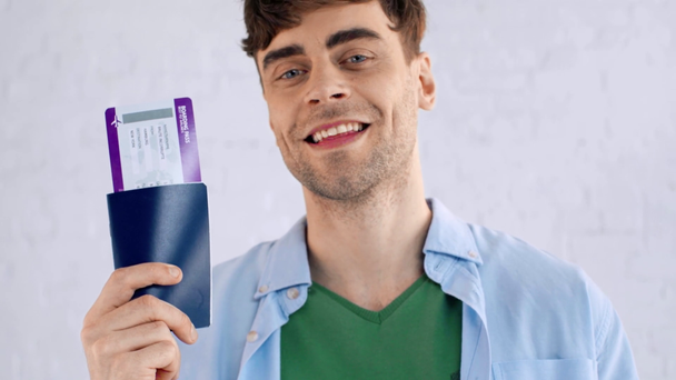 szczęśliwy młody człowiek prezentujący paszport i bilet lotniczy podczas uśmiechu w aparacie - Materiał filmowy, wideo