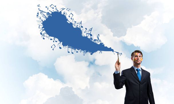 Hombre de negocios exitoso en traje negro haciendo gestos y sonriendo mientras está de pie con salpicadura musical de color azul contra el paisaje nublado vista sobre el fondo
. - Foto, Imagen