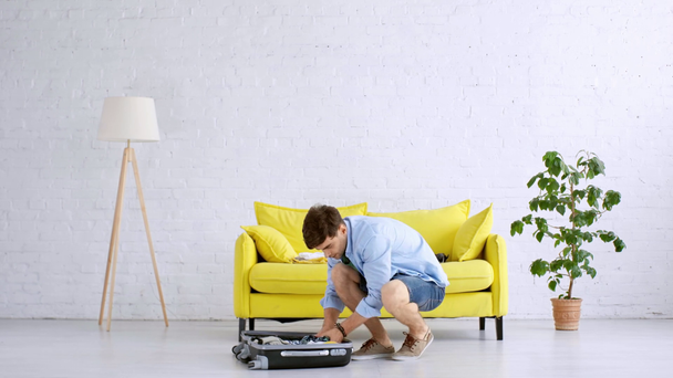 молодой человек распаковывает чемодан на полу и кладет одежду на желтый диван
 - Кадры, видео