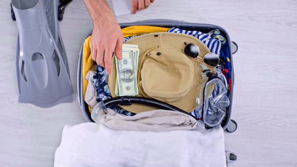 cropped view of man packing summer clothes, swimming accessories, money, passport an air ticket in suitcase on floor at home - Footage, Video