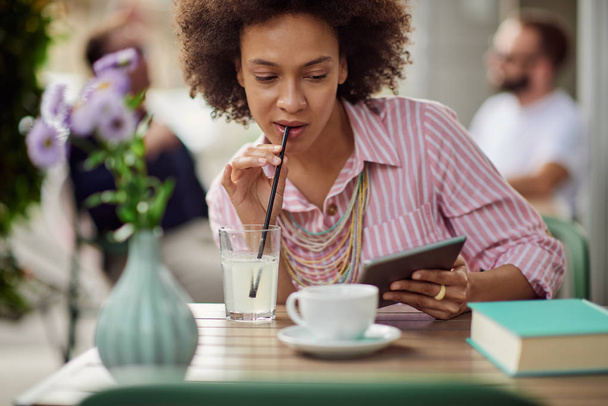 Великолепная женщина смешанной расы в розовом полосатом платье пьет лимонад и использует планшет, сидя в кафе
. - Фото, изображение