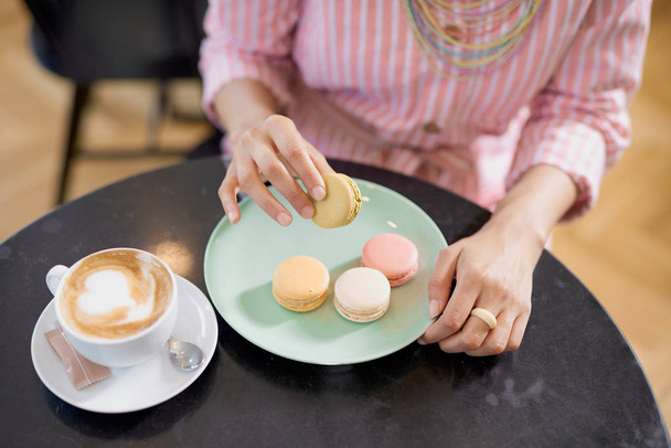 Vista superior de la mujer de raza mixta en vestido de rayas rosadas sosteniendo la galleta mientras está sentado en la pastelería. En la mesa hay café y plato con galletas
. - Foto, imagen