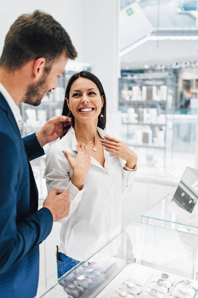 Beautiful couple enjoying in shopping at modern jewelry store. Young woman try it out gorgeous necklace and earrings. - Foto, Bild