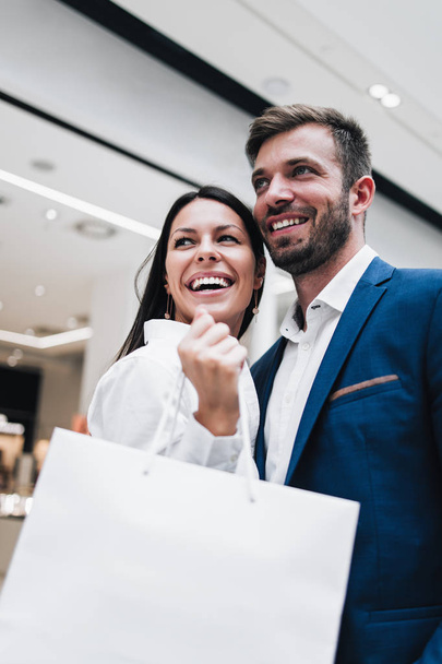 Beautiful couple enjoying in shopping mall or center. They standing in front of huge modern store, smiling and looking at side while holding shopping bags. Shoot from below. - Foto, immagini