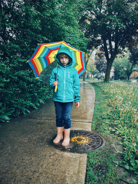 Valkoihoinen tyttö seisoo lätäkössä sateen aikana sateenkaaren alla kevään kesäpäivänä ulkona. Onnellinen lapsuuden aito elämäntapa. Sävytetty retro vintage hipster elokuva suodattimet
. - Valokuva, kuva