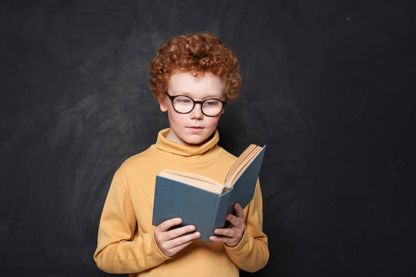 Child reading a book on blackboard background. Cute little boy  - Photo, Image
