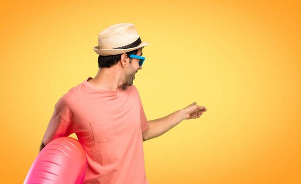 Mann mit Hut und Sonnenbrille im Sommerurlaub zeigt mit dem Zeigefinger zurück und präsentiert ein Produkt auf orangefarbenem Hintergrund - Foto, Bild