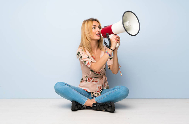 Young blonde woman sitting on the floor shouting through a megaphone - Фото, изображение