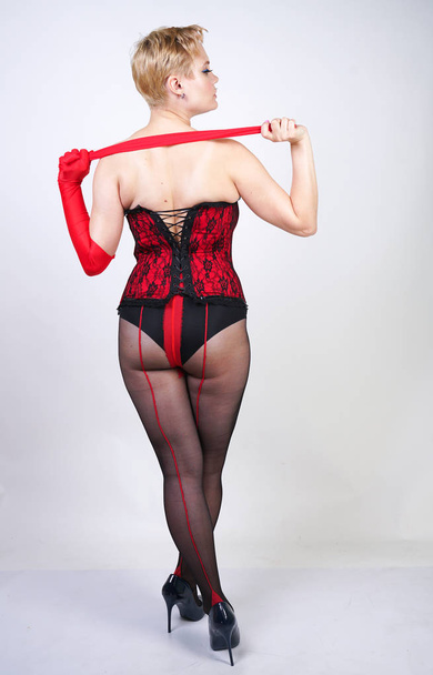 sexy plus size woman with short hair in a red lace bodice and sheer classic black pantyhose with a seam posing on a white background in the Studio - Foto, imagen