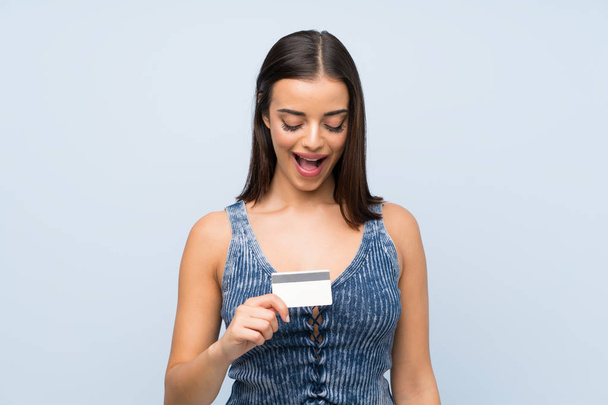 Bir kredi kartı tutan izole mavi duvar üzerinde genç kadın - Fotoğraf, Görsel