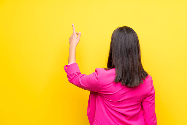 Молодая женщина на изолированном желтом фоне указывает назад указательным пальцем
 - Фото, изображение