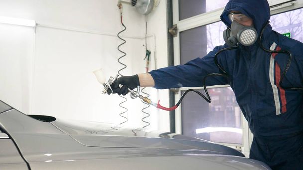 Professional in working uniform with spray guns in his hands and a respirator in protective glasses puts paint (ceramics) on the car on different parts of the car, flashlight with a folder in his hands checks a car engine for serviceability. Concept  - Foto, Bild