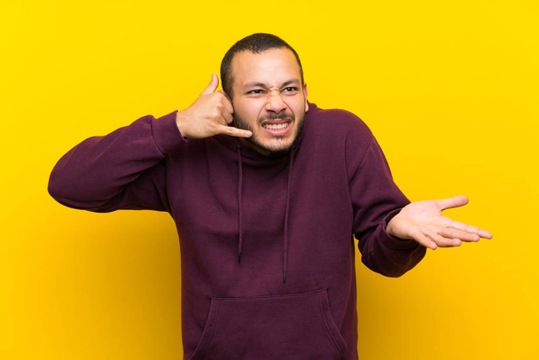 Kolumbijski człowiek z bluzką na żółtym ścianie podejmowania gest telefonu i wątkających - Zdjęcie, obraz