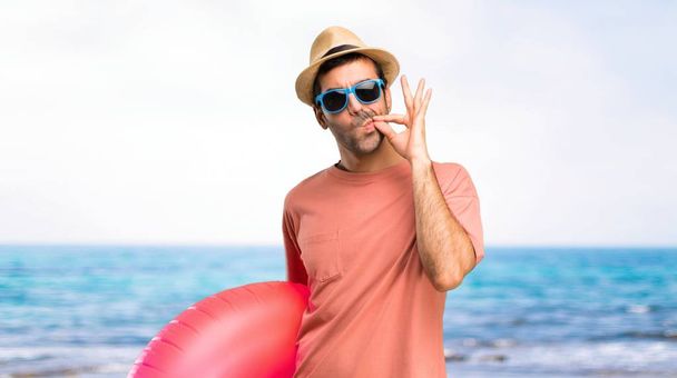 Чоловік з капелюхом і сонцезахисними окулярами на літніх канікулах показує знак закриття рота і тиші жест робить як закриття рота блискавкою на пляжі
 - Фото, зображення
