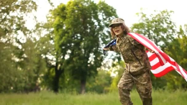 Teenager in camouflage at a picnic in honor of the celebration of US Independence Day. A child runs across the meadow with a large national flag - Video, Çekim