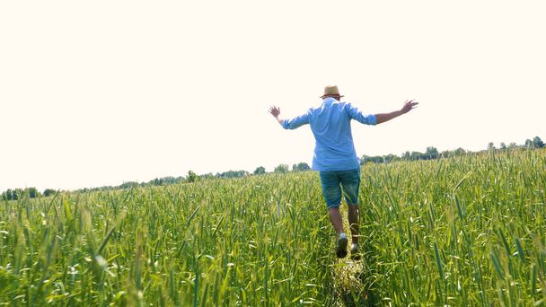 Portret młodego faceta (człowieka) w mundurze roboczym i Kapelusz Słomkowy w środku pola wokół pszenicy i siana, bieganie wokół niego i sprawdzanie pszenicy do dojrzałości, pole ciepła w koszulce i słomy kapelusz oddycha na świeżym powietrzu i czuje się swobodnie.  - Zdjęcie, obraz