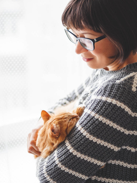 Cute ginger cat dozing on woman hands. Smiling woman in grey knitted sweater stroking her fluffy pet. Cozy home. - Fotografie, Obrázek