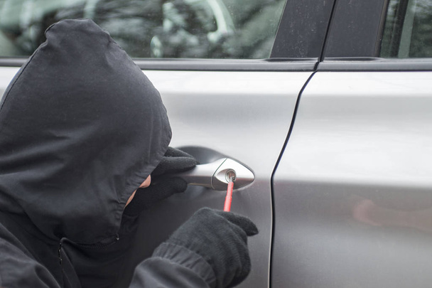 男は車に押し入ろうとして彼の頭の上フード付き黒に身を包んだ。車泥棒は、車の盗難のコンセプト - 写真・画像