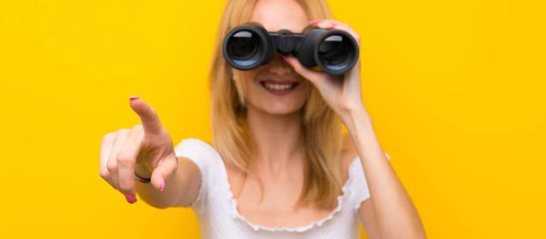 Mujer rubia joven sobre pared amarilla aislada con prismáticos negros
 - Foto, Imagen