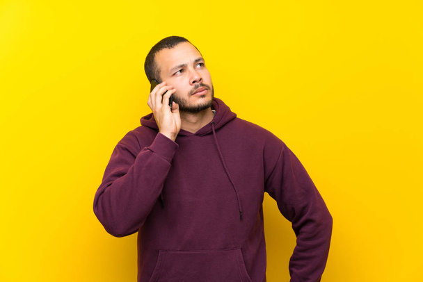 Colombian man with sweatshirt over yellow wall keeping a conversation with the mobile phone - Φωτογραφία, εικόνα