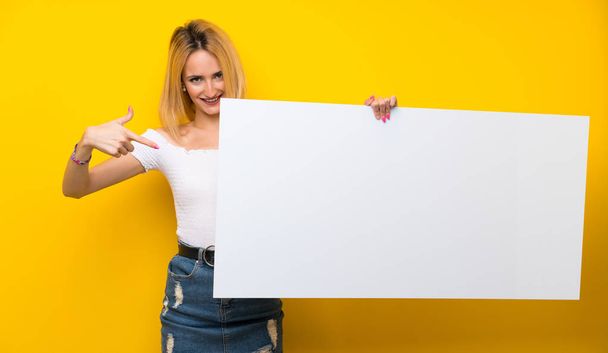Молодая блондинка над изолированной желтой стеной с пустым белым плакатом для вставки концепции
 - Фото, изображение