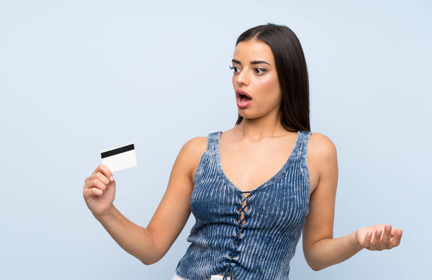Young woman over isolated blue wall holding a credit card - Foto, Imagem