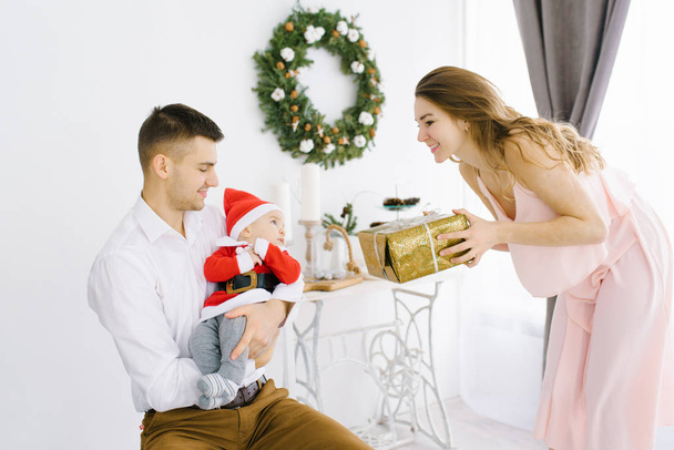 Papa hält den Sohn im Weihnachtsmann-Anzug auf dem Arm, Mama beschenkt das Kind auf dem Hintergrund des Weihnachtsbaums im Wohnzimmer. Weihnachtsurlaub in der Familie. Glück und Menschen - Foto, Bild