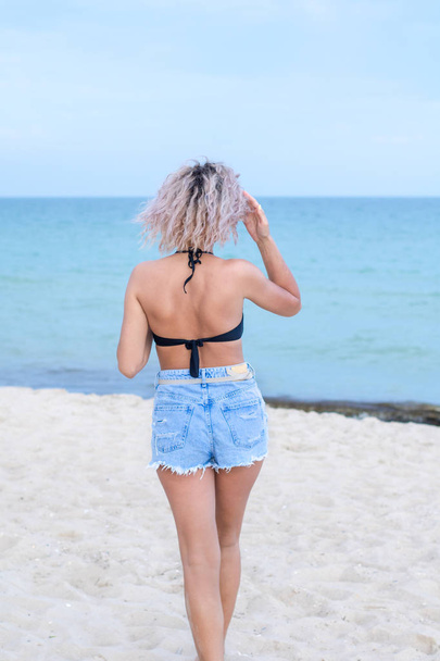 красива молода жінка з кучерявими волоссям в джинсових шортах і купальнику щаслива бути на пляжі біля океану. Океан і трава на фоні
 - Фото, зображення