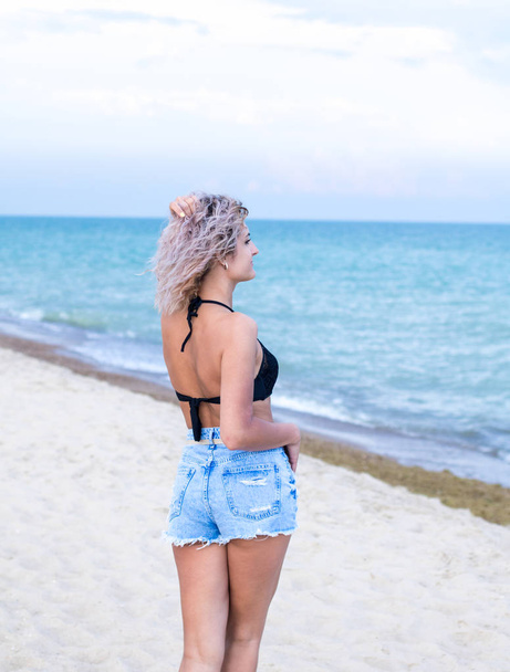 krásná mladá žena s kudrnatými vlasy v džíšových trenýrkách a plavky je šťastná, že je na pláži blízko oceánu. Oceán a tráva na pozadí - Fotografie, Obrázek