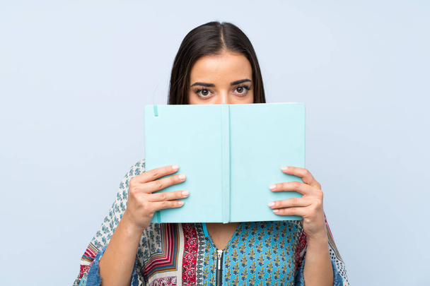 Jonge vrouw over geïsoleerde blauwe muur houden en lezen van een boek - Foto, afbeelding