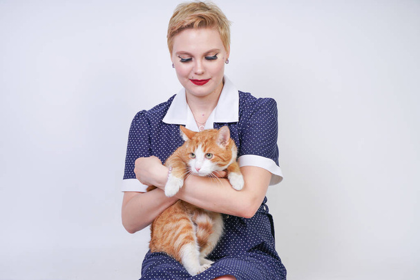 Cute Kobieta rodzaju z krótkimi włosami w Pinup polka dot sukienka trzymając jej ukochanego zwierzaka na białym tle w Studio. plus rozmiar dorosły blond dziewczyna i jej pomarańczowy kot są szczęśliwi razem. - Zdjęcie, obraz