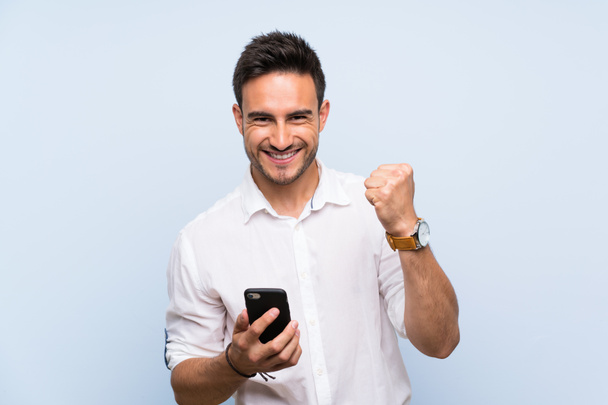 Handsome young man over isolated blue background with phone in victory position - Photo, Image