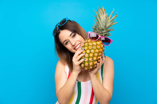 Νεαρή γυναίκα το καλοκαίρι διακοπές πάνω από το μπλε φόντο κρατώντας ένα ανανά με γυαλιά ηλίου - Φωτογραφία, εικόνα